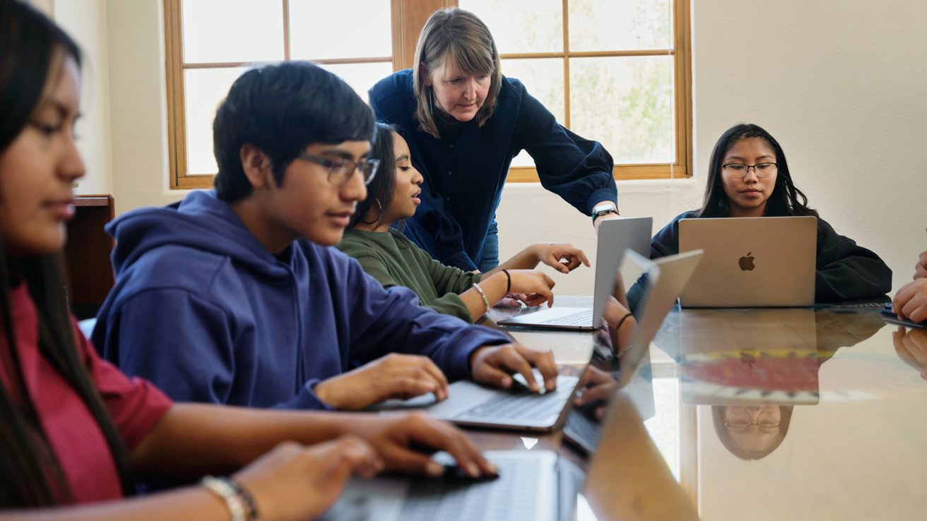 Teacher helps students working on laptops around a table in a classroom with large windows in the background.