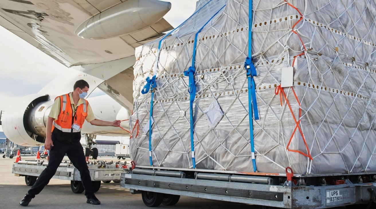 Airport worker in a safety vest and mask unloads a large, netted cargo from an aircraft onto a rolling platform.