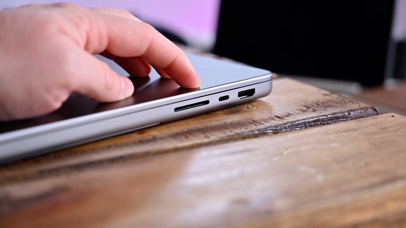 Hand touching a closed laptop on a wooden surface, focusing on the side ports including USB and HDMI slots.