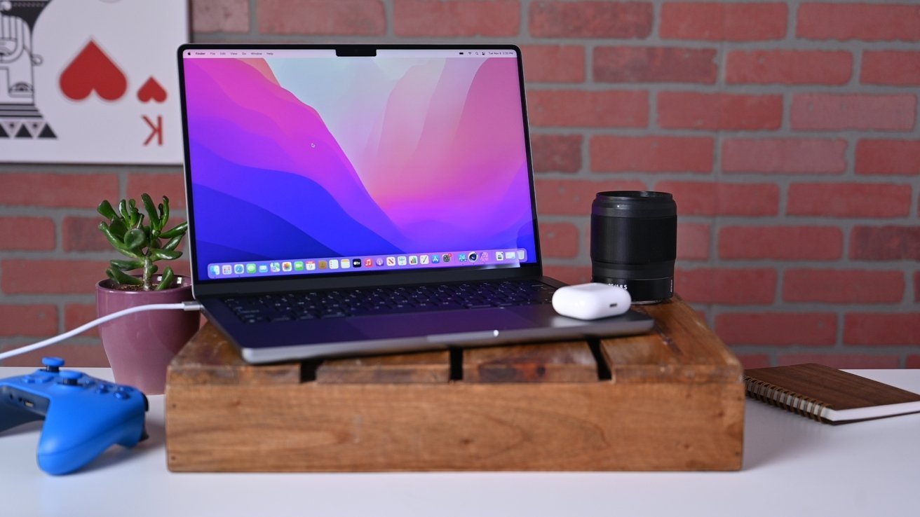 A laptop with a colorful screen sits on a wooden stand, surrounded by a camera lens, earbuds, a notebook, a game controller, and a small plant. Brick background.