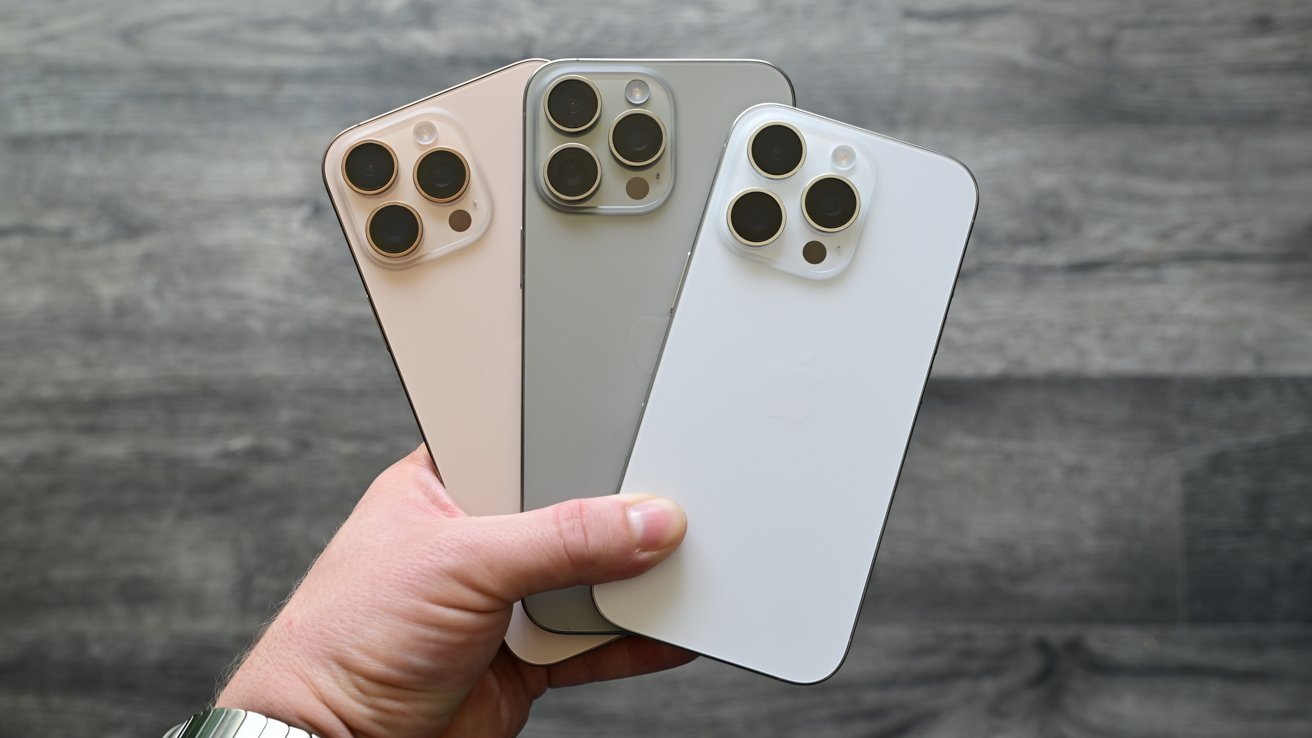 A hand holds three smartphones with triple cameras and different colors, against a textured gray background.