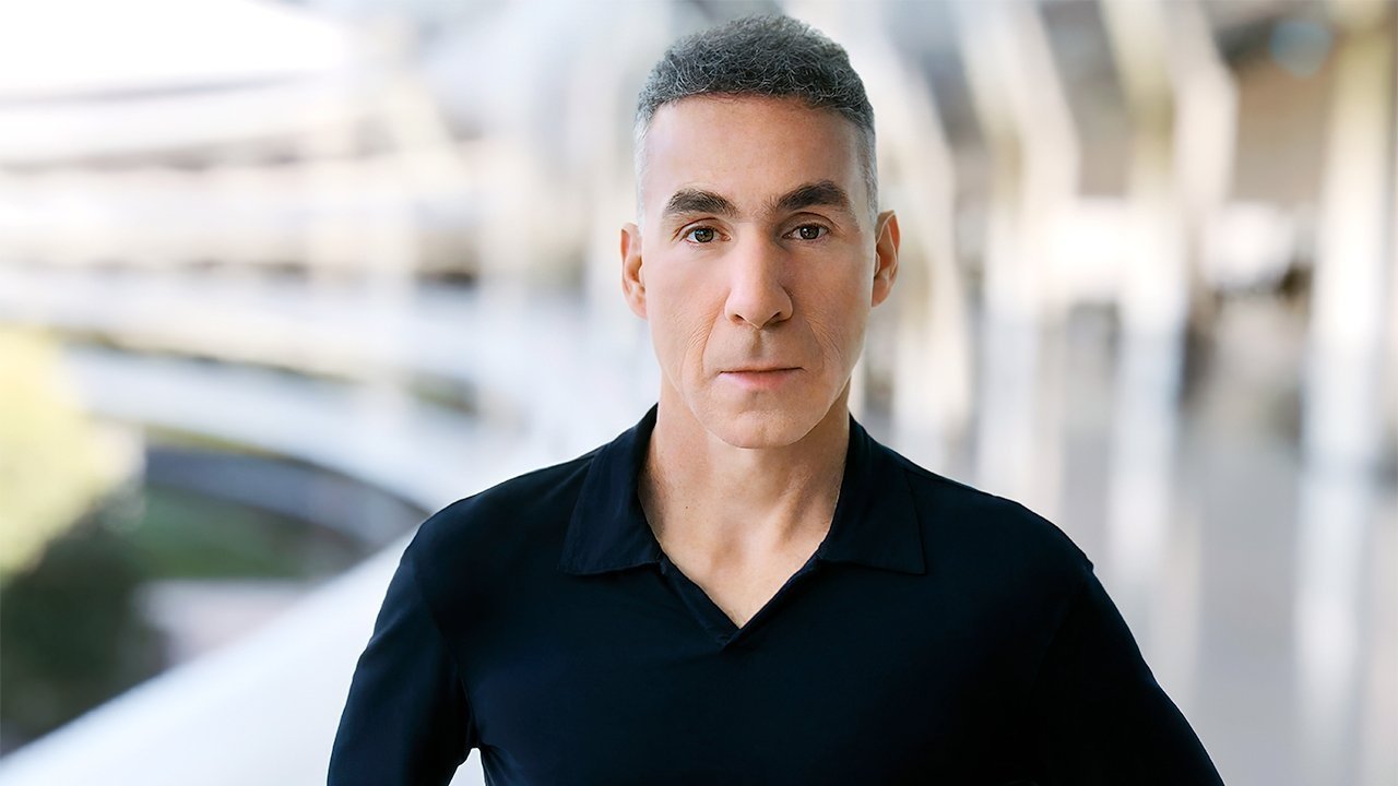 A serious-looking man with short hair wearing a dark shirt stands against a blurred modern architectural background.