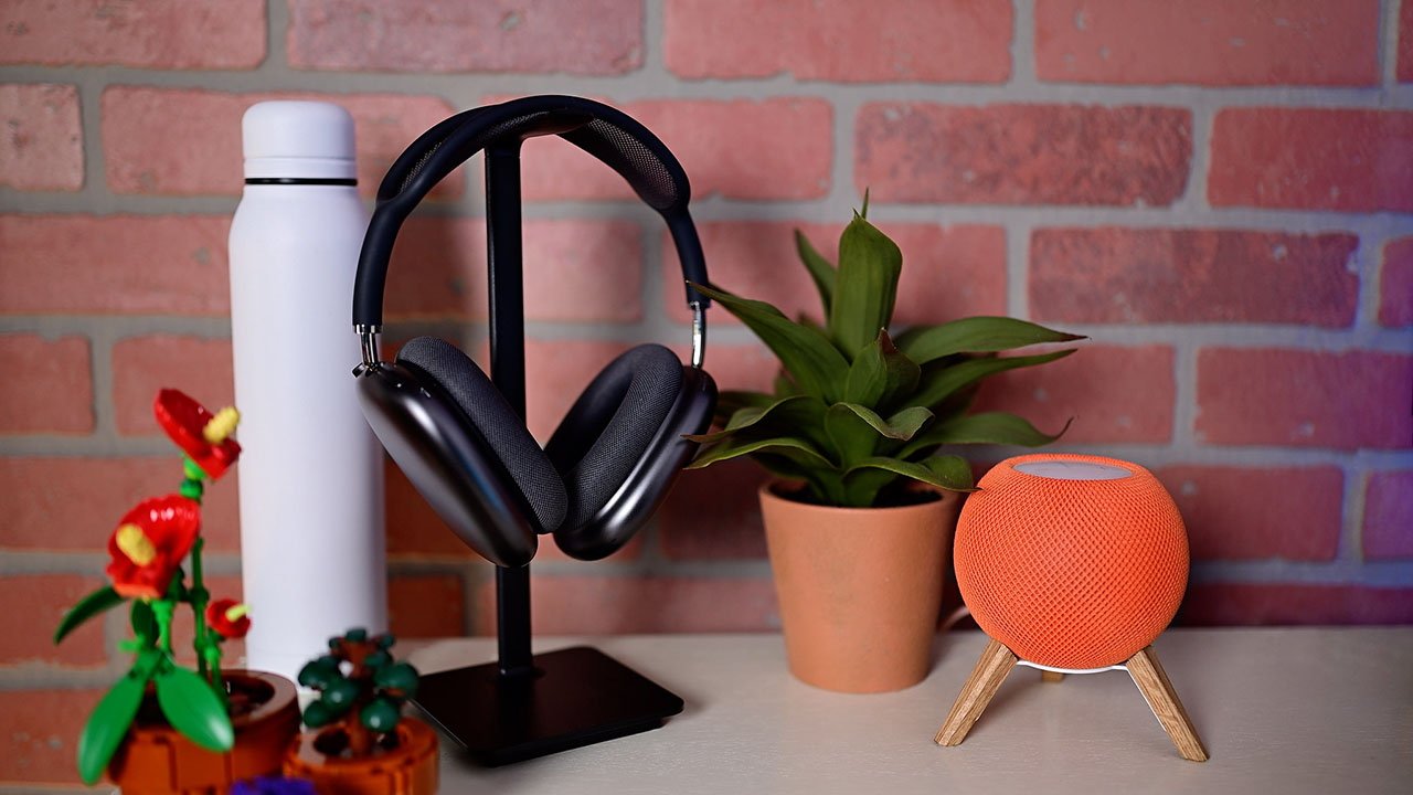 AirPods Max en un soporte, botella blanca, planta en maceta, mini altavoz HomePod naranja y flores de juguete en una mesa contra un fondo de pared de ladrillo.