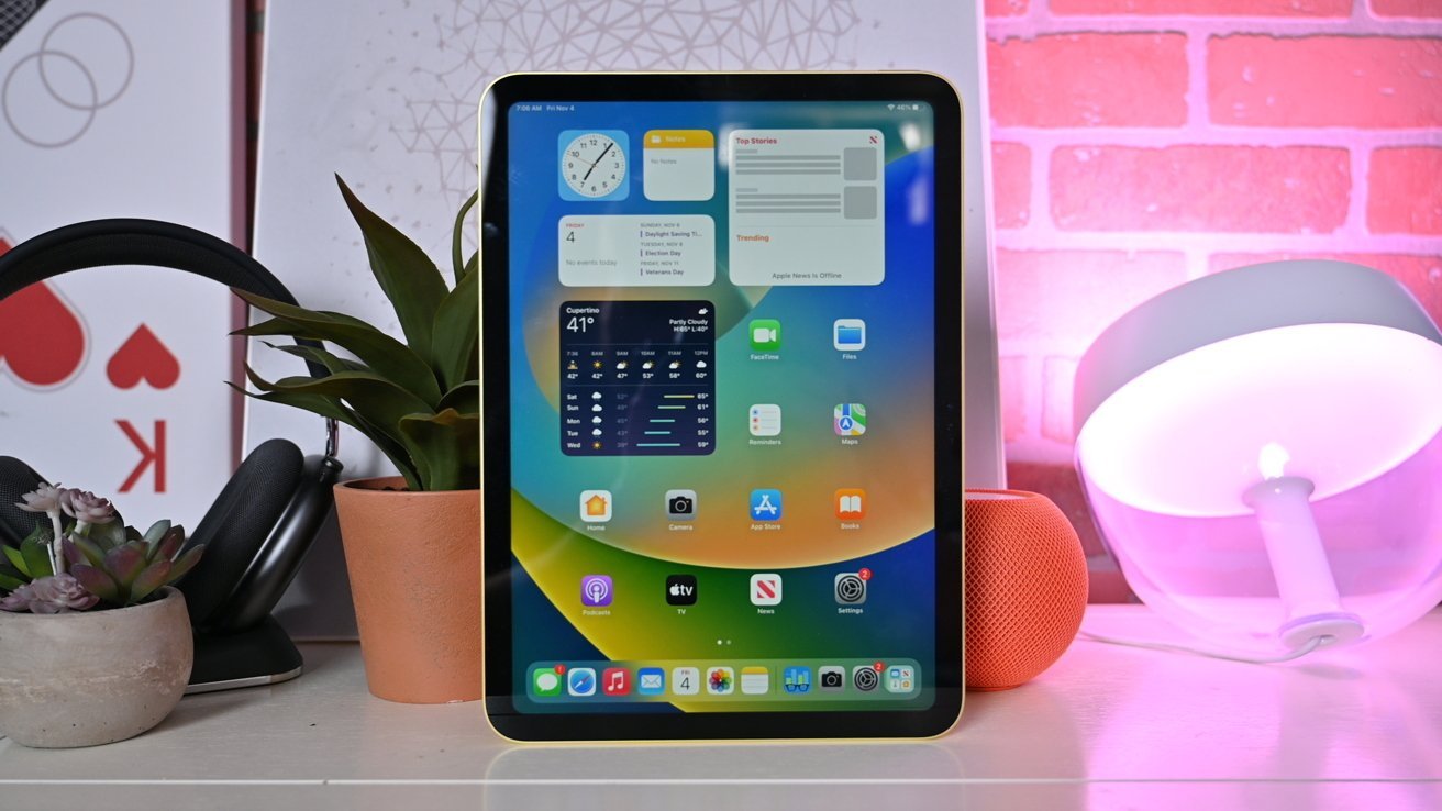 A 10th generation iPad displaying weather and apps, surrounded by plants, headphones, and a table lamp with pink lighting.