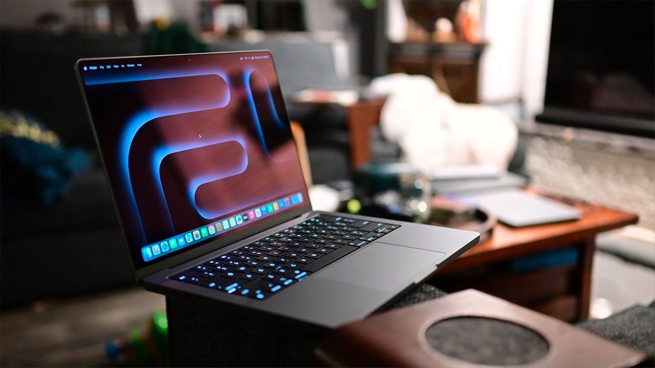 Un Apple MacBook Pro de 14 pulgadas en color gris espacial con un teclado iluminado y un salvapantallas abstracto está colocado sobre un sofá en una habitación con poca luz.