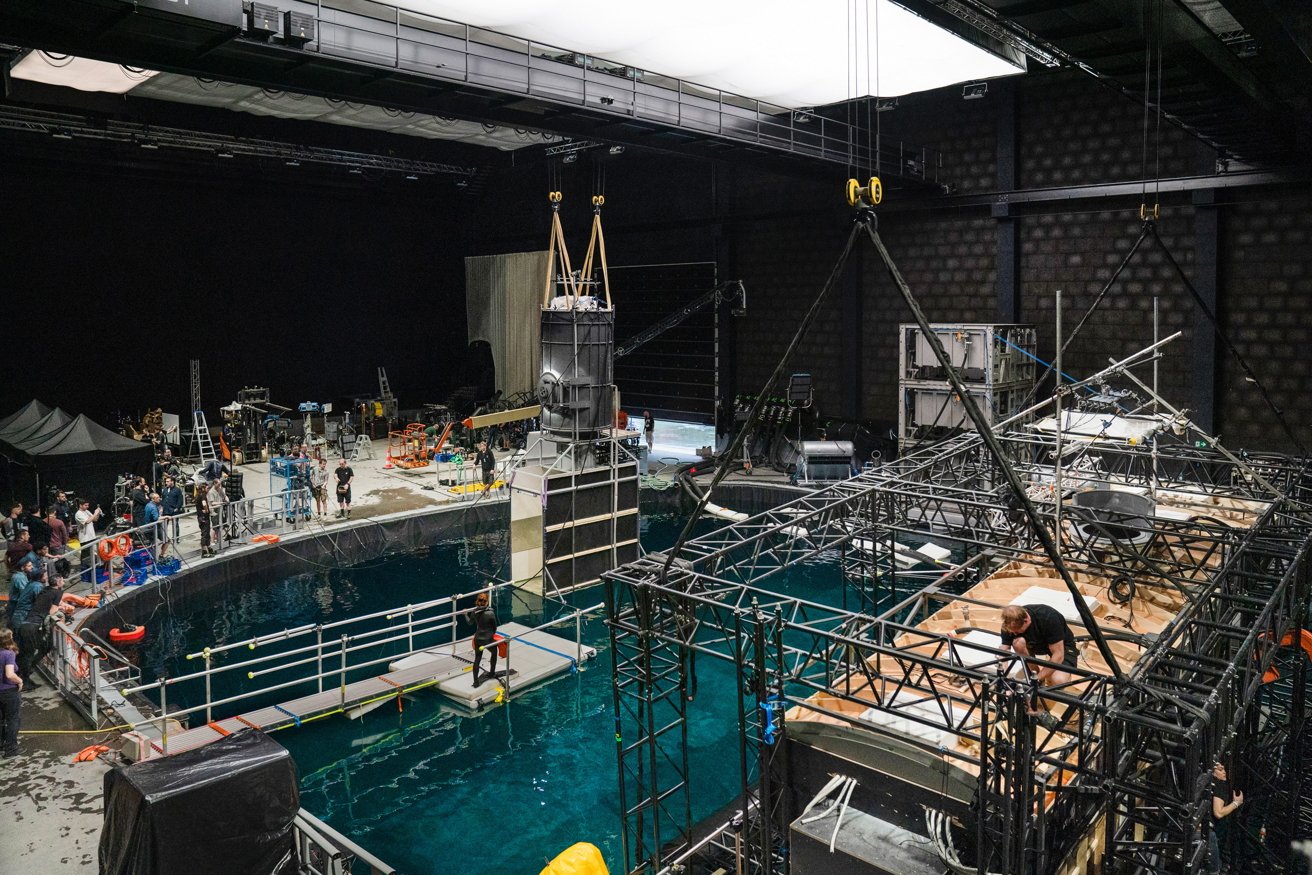 Indoor film set with a large water tank, scaffolding, and various equipment. Crew members work around the area, some near the tank and others on the scaffolding.