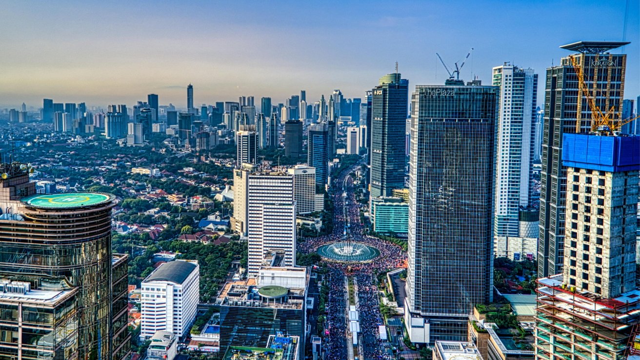 Aerial view of a bustling cityscape featuring skyscrapers, dense buildings, and a crowded roundabout with a fountain at the center, set under a clear blue sky.