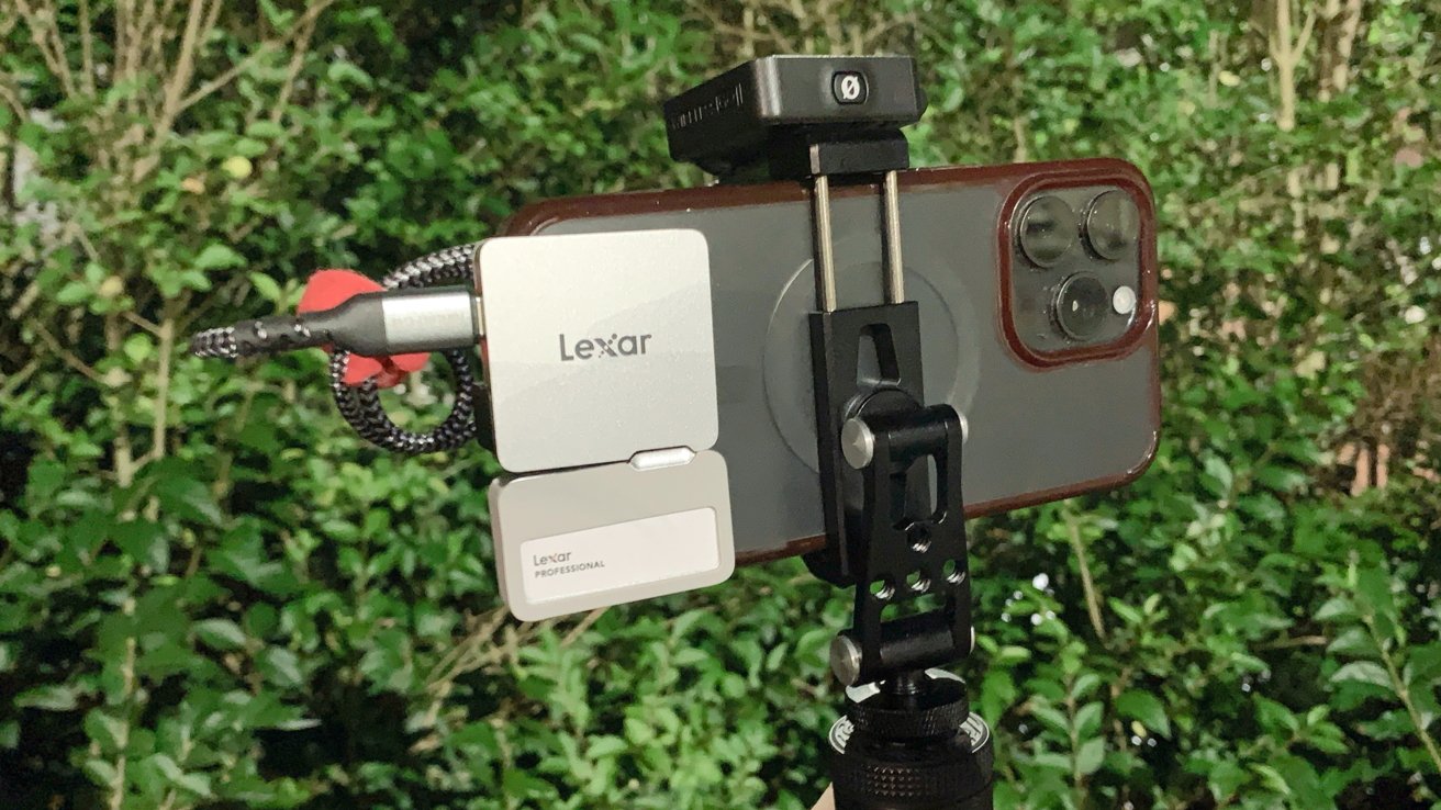 A smartphone is mounted on a tripod with an attached Lexar device and microphone against a backdrop of lush green foliage.