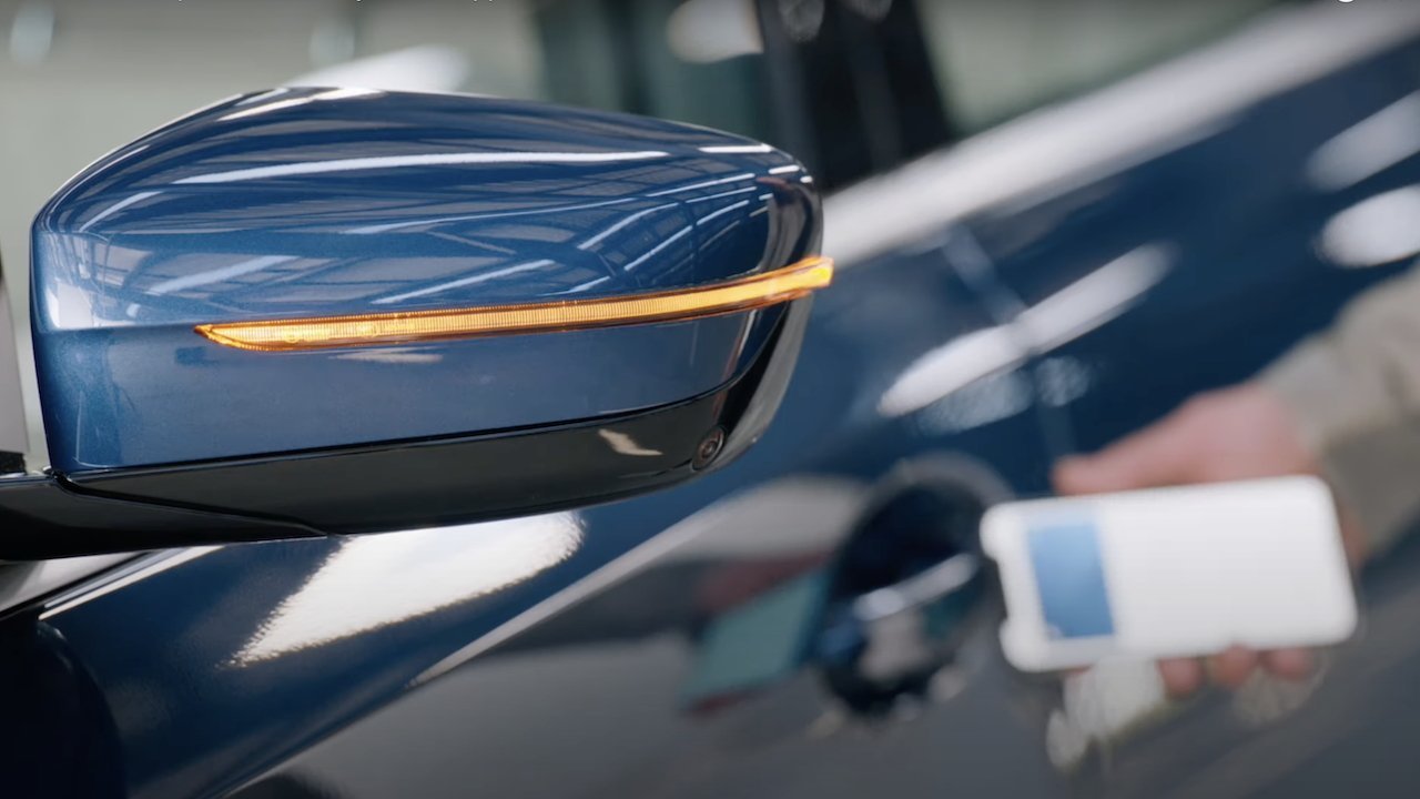 Blue car side mirror with an orange indicator light, a hand holding a smartphone near a door handle in the background.