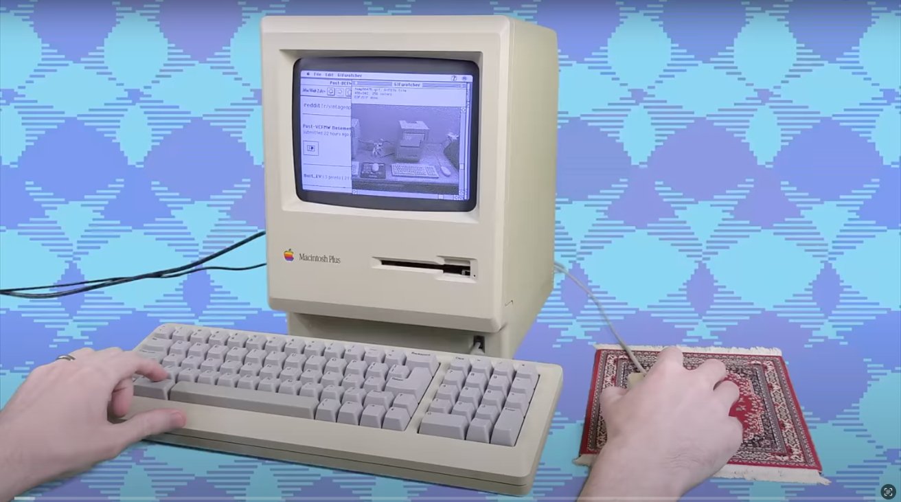 A person uses a retro Macintosh computer with a keyboard and small mouse pad on a patterned blue background.