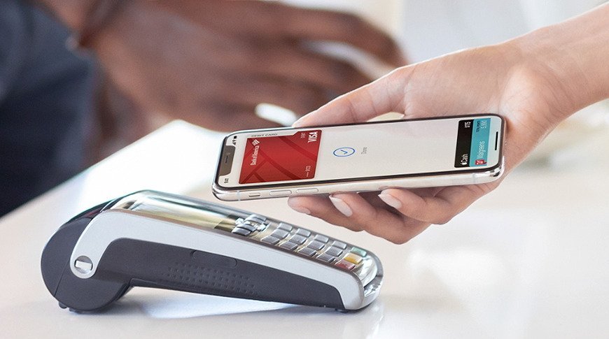 A hand holds a smartphone near a payment terminal, showing a contactless payment being made with a digital wallet app displayed on the screen.