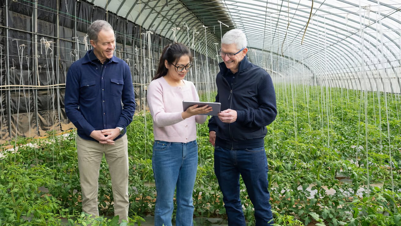 Three people stand in a greenhouse, looking at a tablet. Lush green plants surround them under a curved, transparent roof.