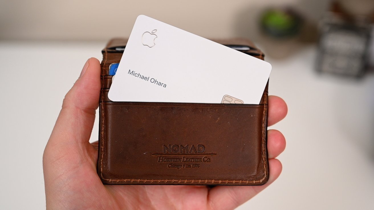 Hand holding a brown leather wallet with a partially visible white card, showing the Apple logo and text.