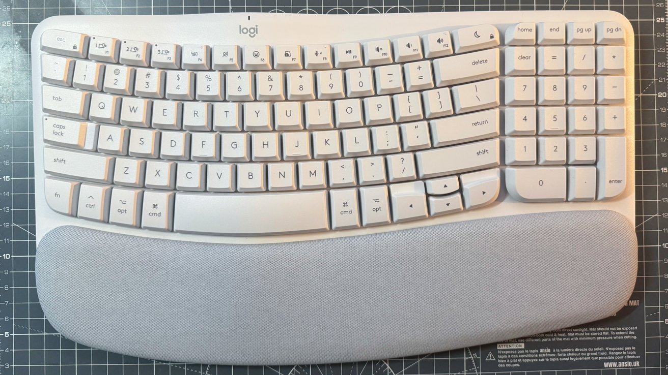 Keyboard putih ergonomis dengan tombol melengkung dan sandaran tangan abu-abu, terletak pada permukaan berpola kotak.