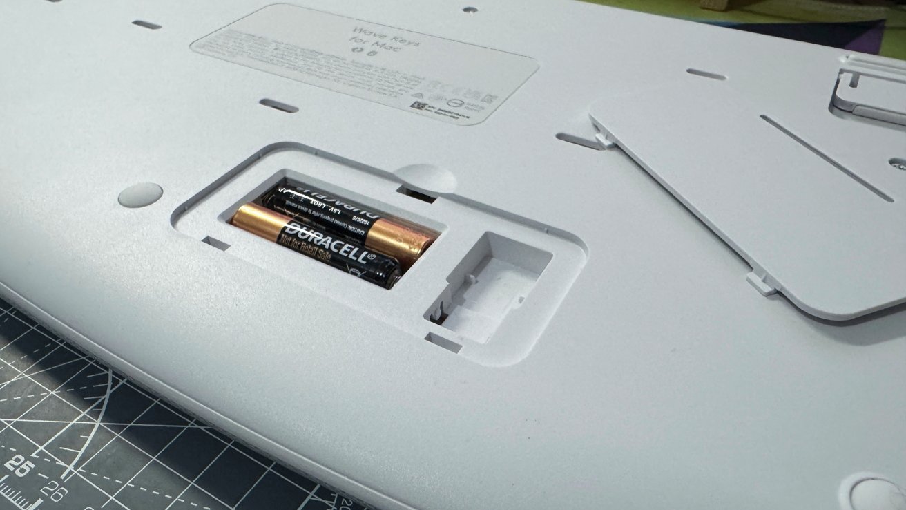 A white electronic device with an open battery compartment showing two AA batteries lies on a cutting mat.