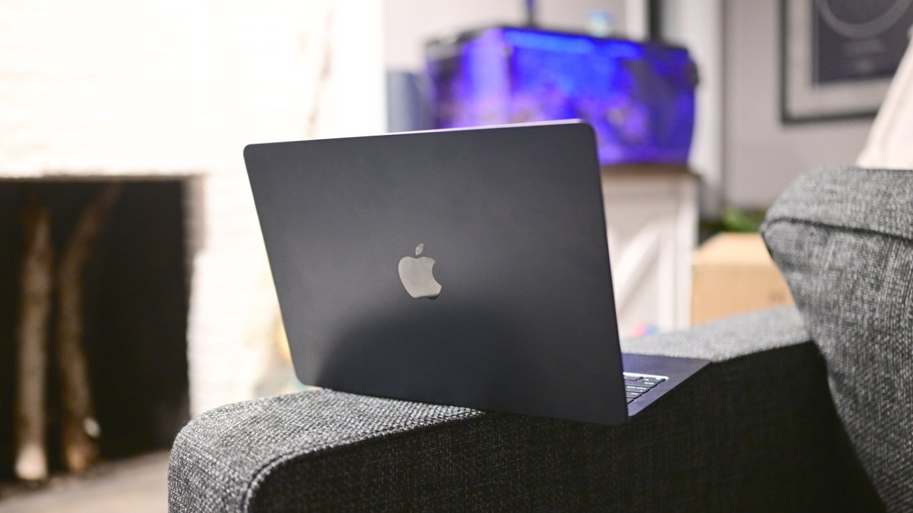 A laptop with an Apple logo sits on a gray couch armrest, with a blue-lit aquarium in the background.