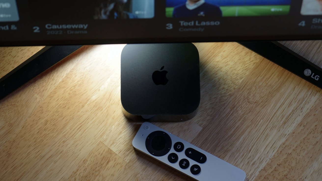 A remote control lies beside a black Apple TV unit on a wooden surface, with part of an LG monitor visible above.