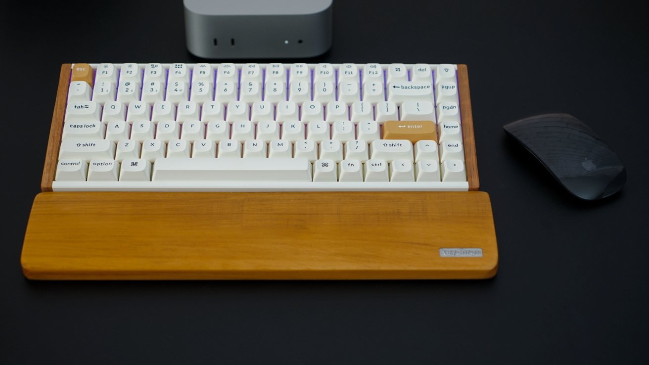 Mechanical keyboard with wooden wrist rest, adjacent to a wireless mouse on a dark surface.