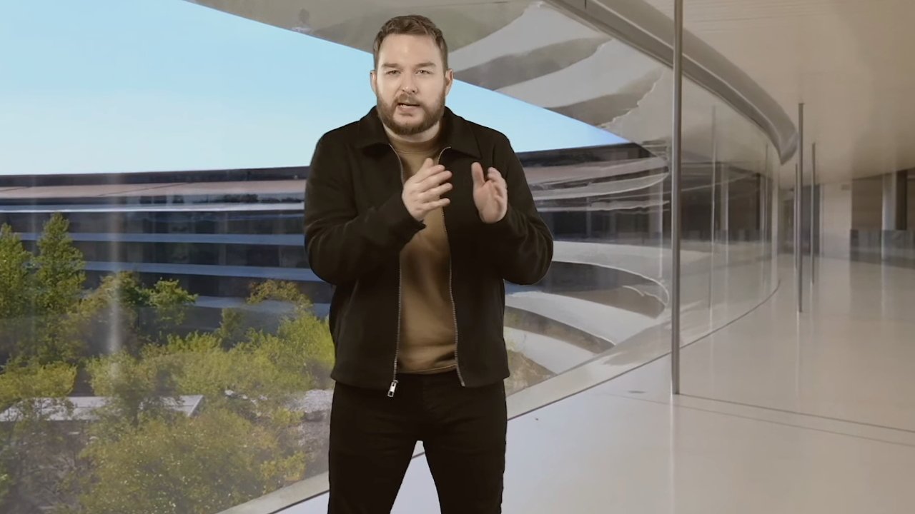 Man in a brown shirt and black jacket stands gesturing in a modern glass-walled building with shrubbery and a blue sky visible outside.