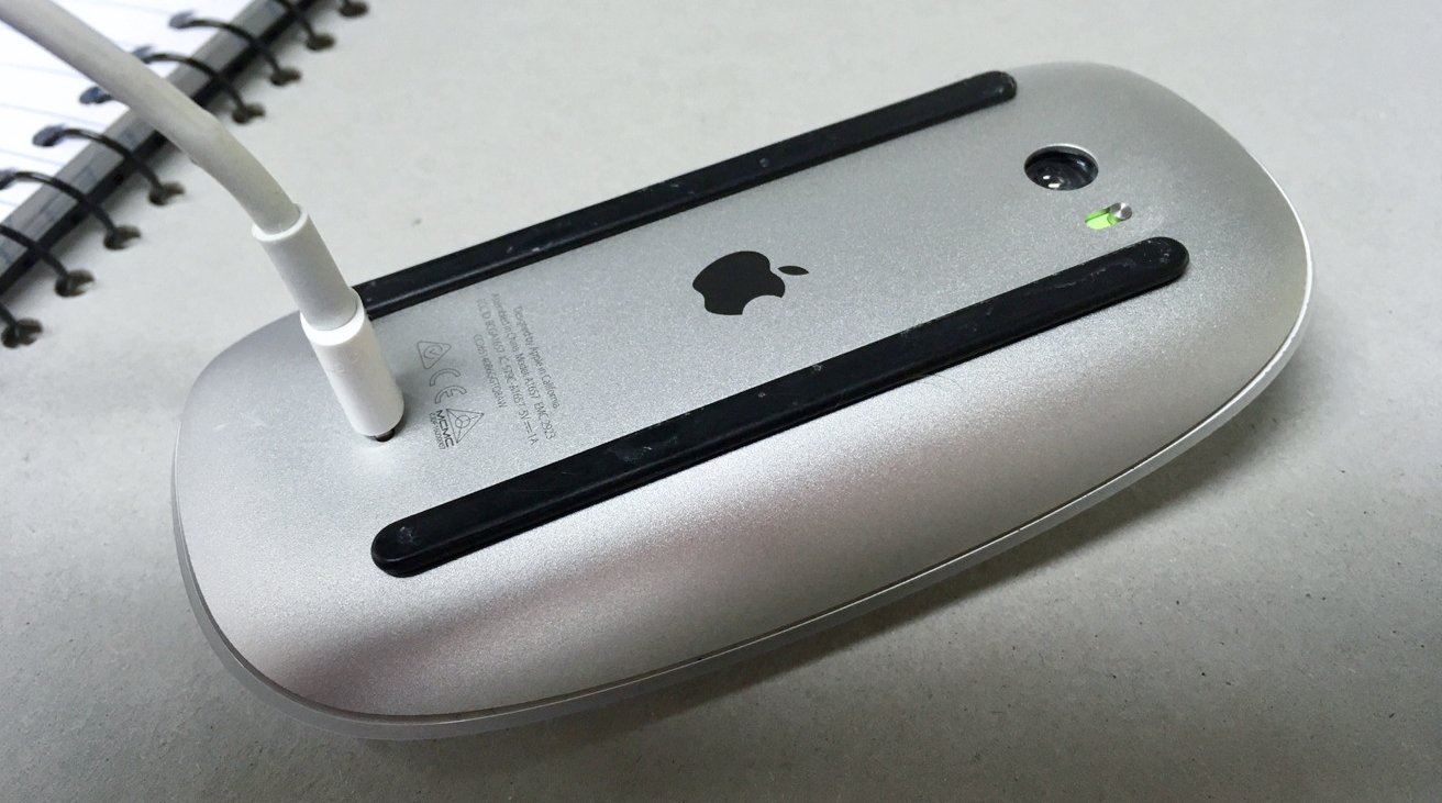 The underside of a silver computer mouse with a charging cable connected, black grip strips, and a partially visible notebook spiral.