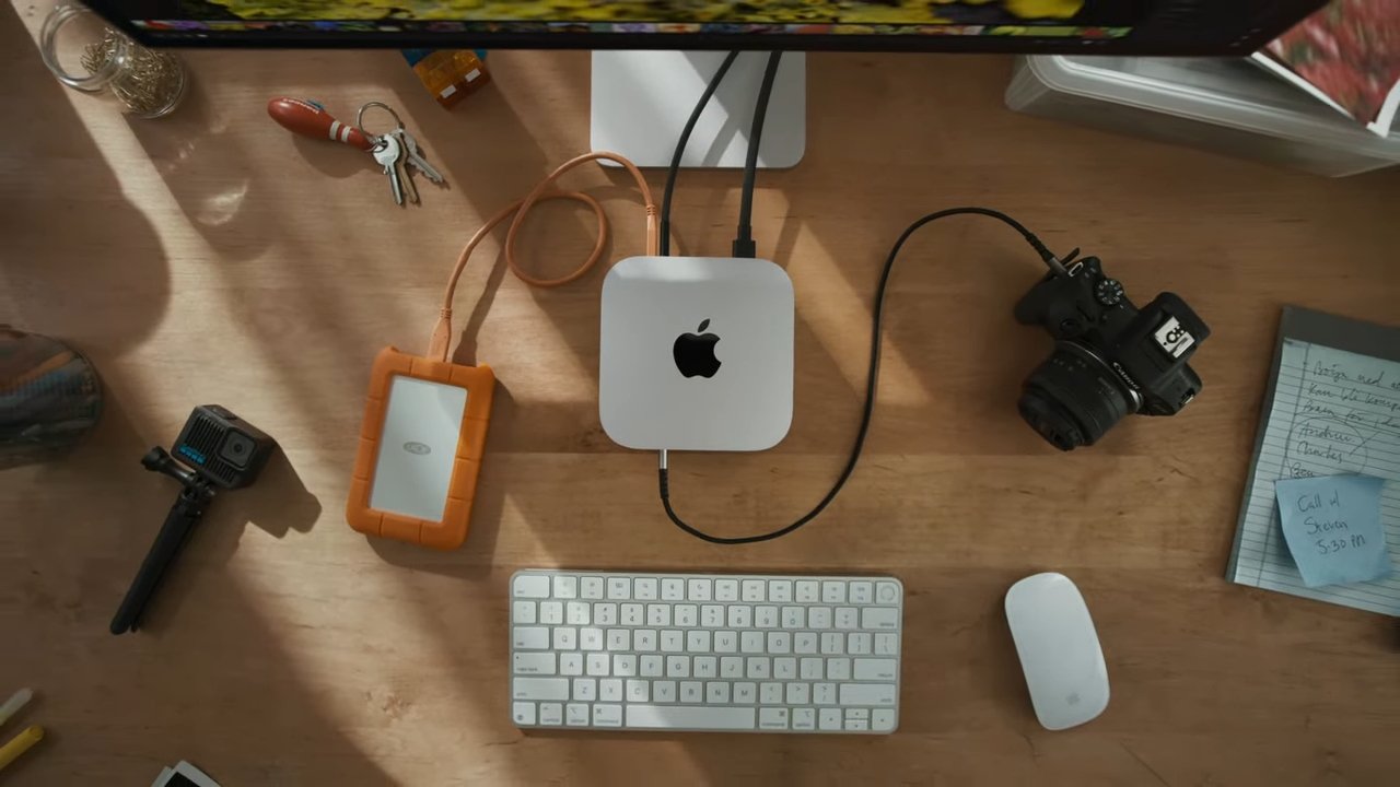 Wooden desk with a computer, keyboard, mouse, camera, orange hard drive, keys, notes, and a GoPro.