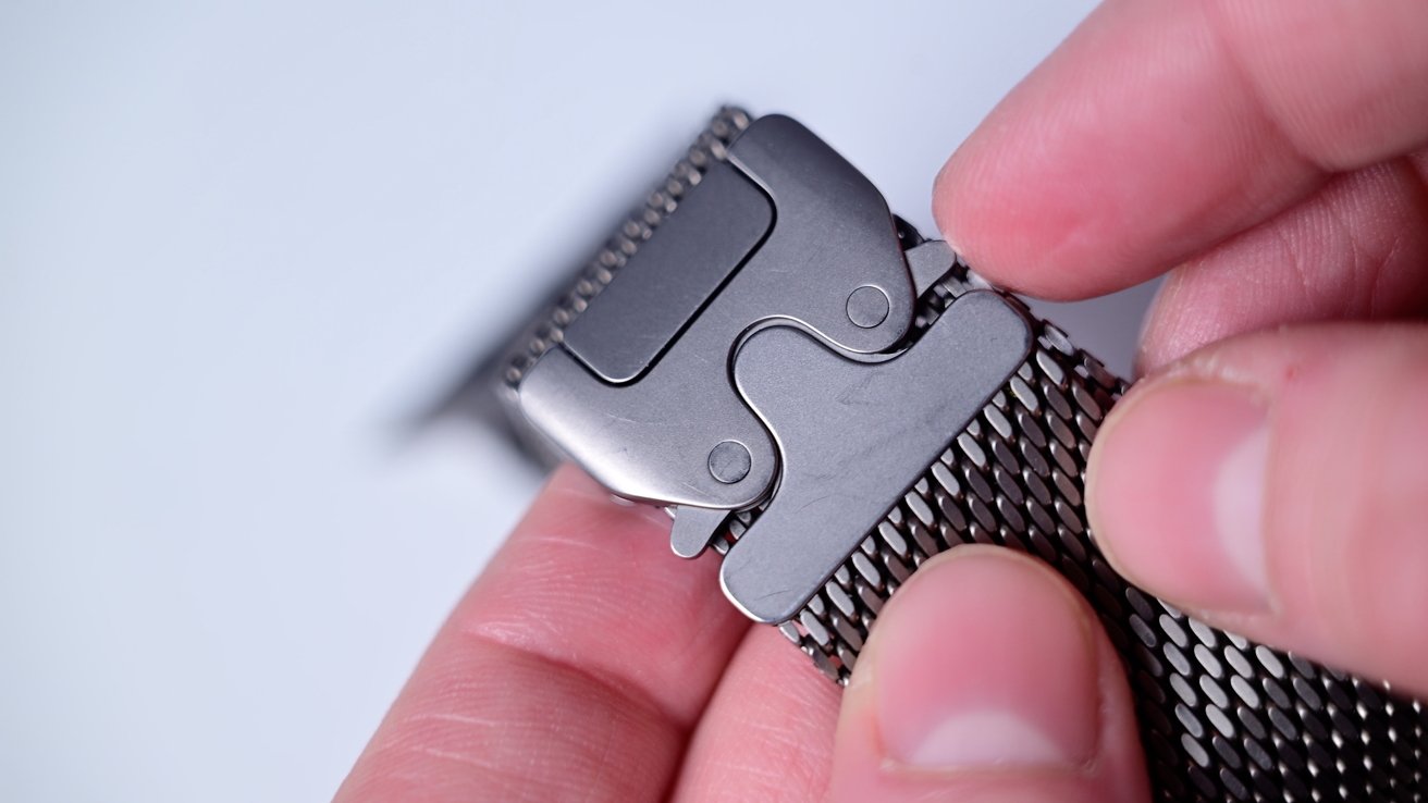 Hands adjust a silver metal lock on a mesh clock band against a regular background.