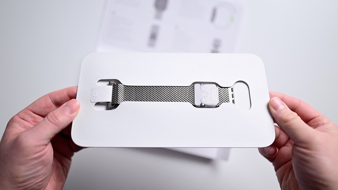 Hands that hold white packaging containing a stainless steel clock band inside. Background shows paper documents outside the focus.