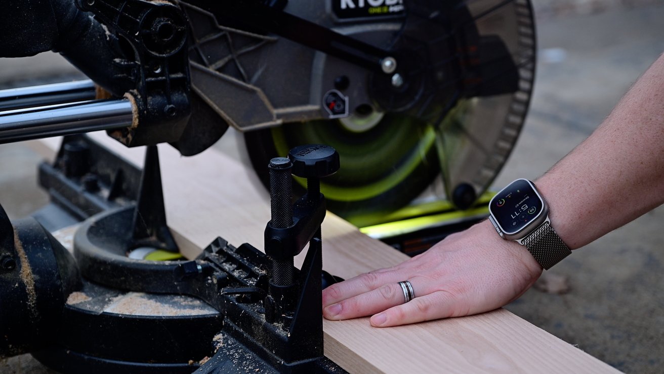 The person's hand wearing a smart trip and leads a piece of wood through a miter looked with a rotating blade.