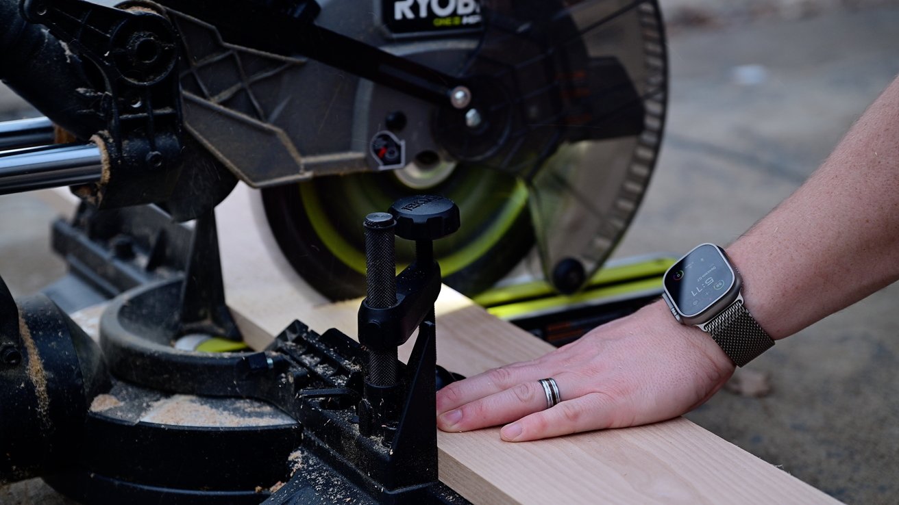 Hand wearing a smartwatch, resting on a piece of wood being cut with a circular saw. Sawdust scattered around.