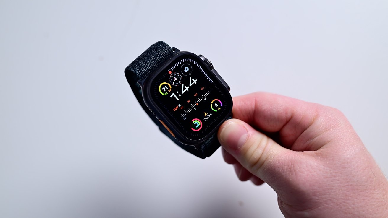 A hand holds a smartwatch displaying colorful fitness rings and the time 7:44 against a plain background.