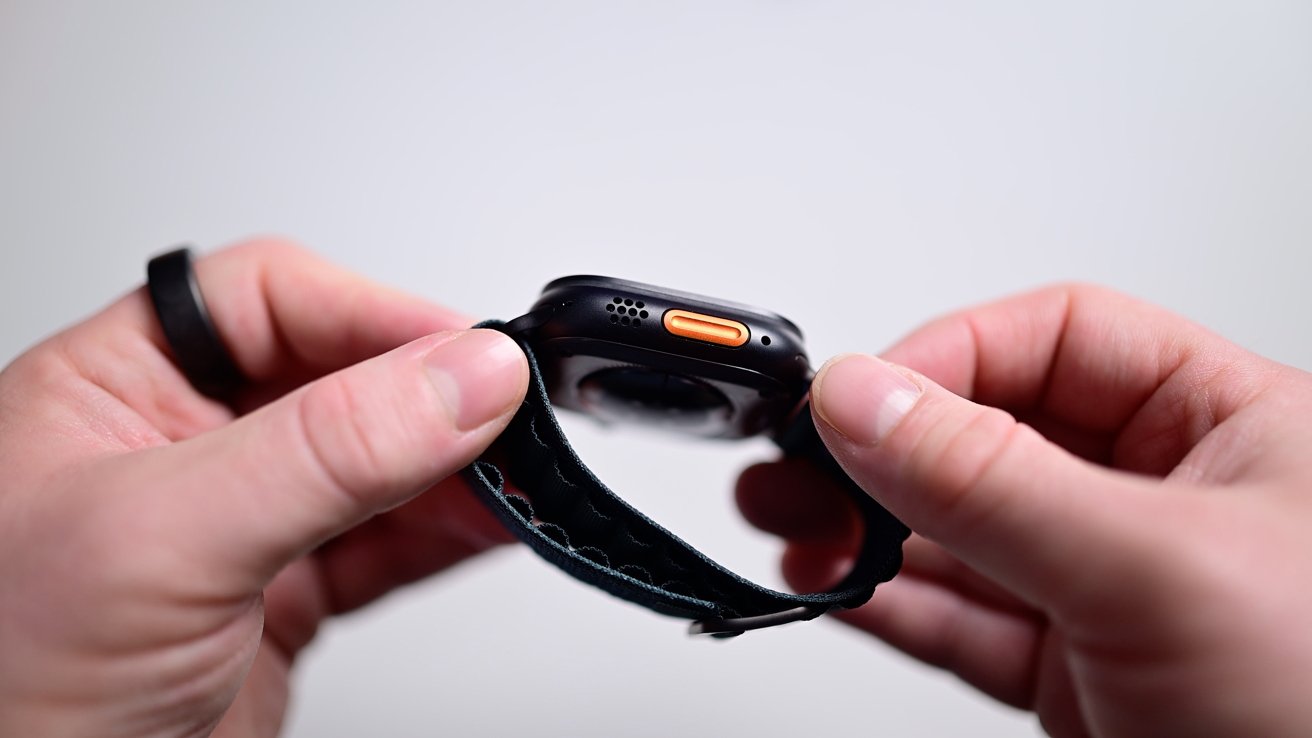 Hands holding a smartwatch with a black band, focusing on an orange button on the side.