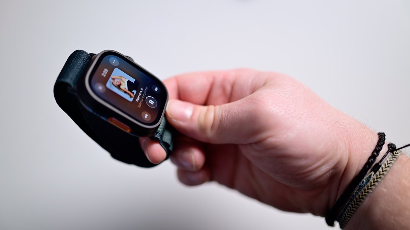 Hand holding a smartwatch displaying a music player with a song paused, featuring artist photo and playback controls.