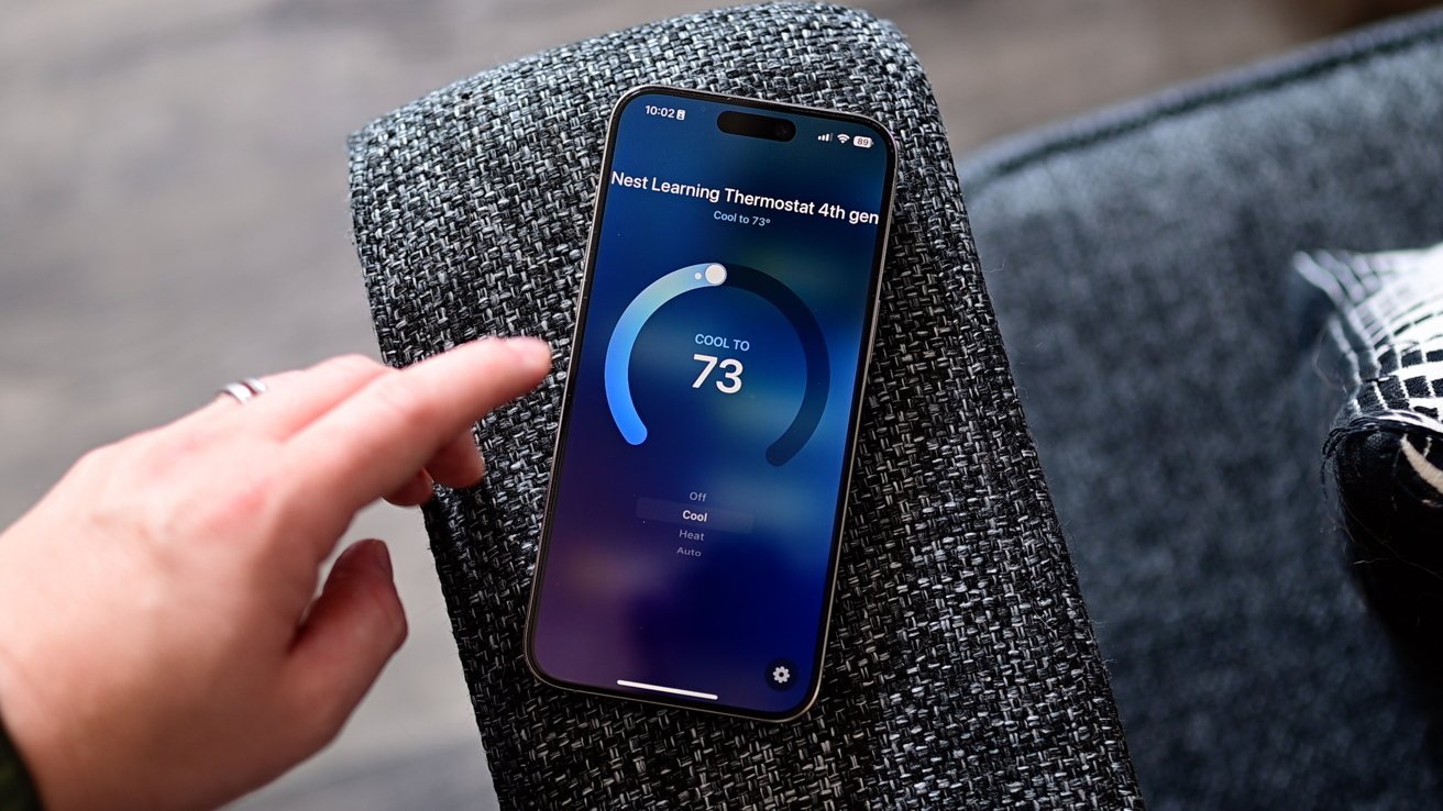 A smartphone on a fabric surface displays a Nest Learning Thermostat app, set to cool at 73 degrees, with a hand approaching the screen.