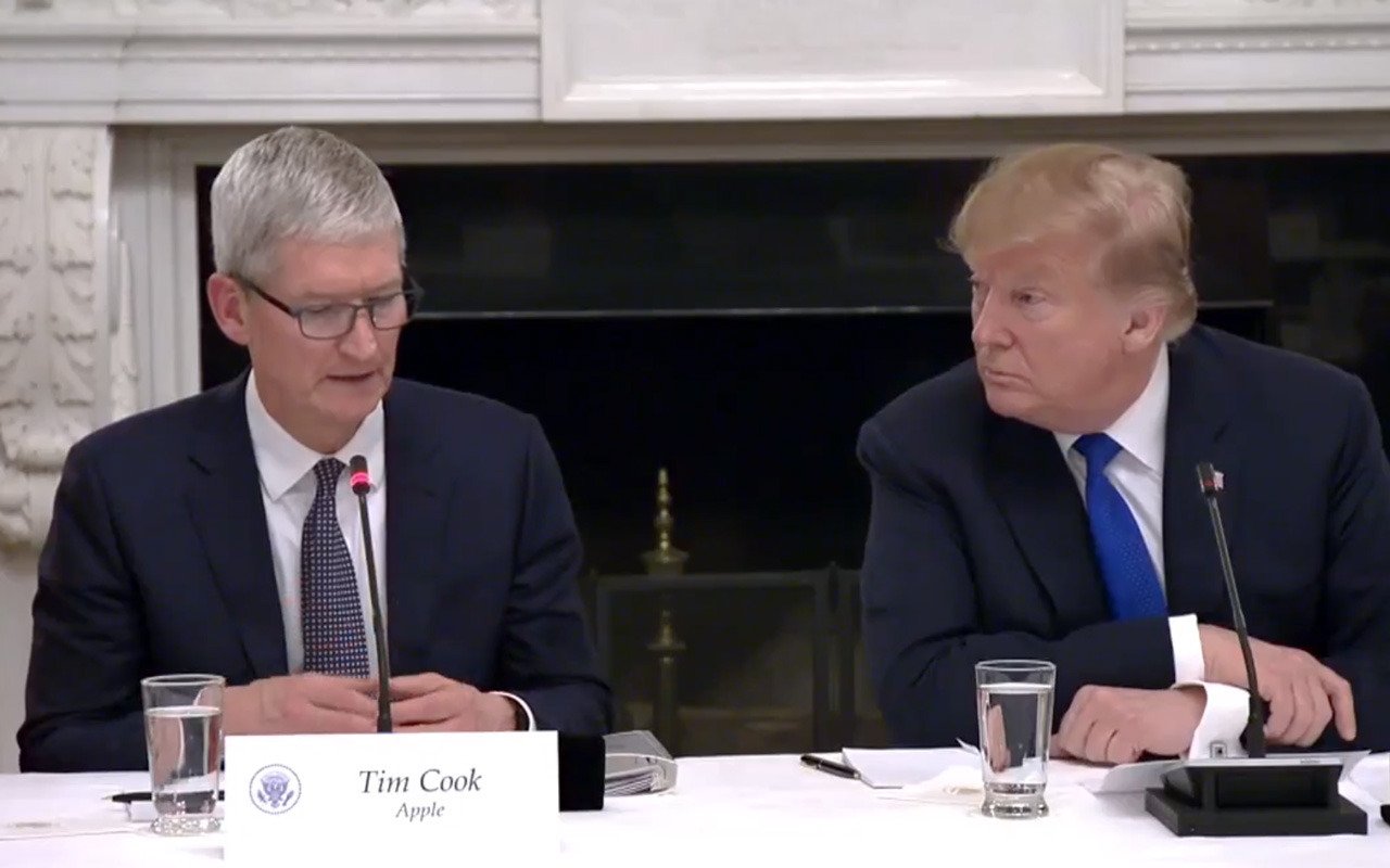 Two men in suits sitting at a roundtable meeting, one speaking into a microphone with a nameplate in front of him, the other listening attentively.