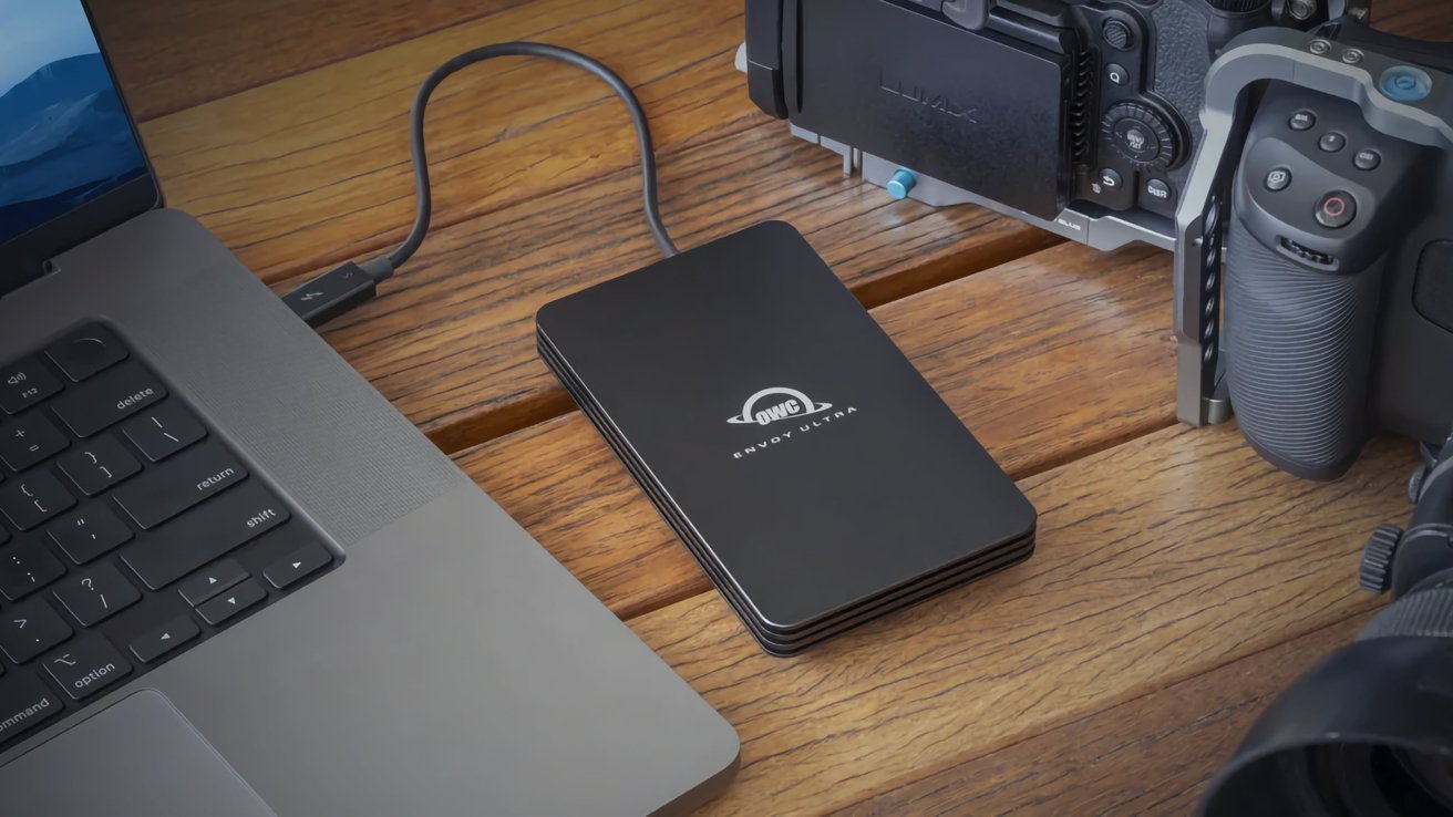 A black external hard drive labeled OWC Envoy Ultra connected to a laptop, placed on a wooden table with a camera nearby.