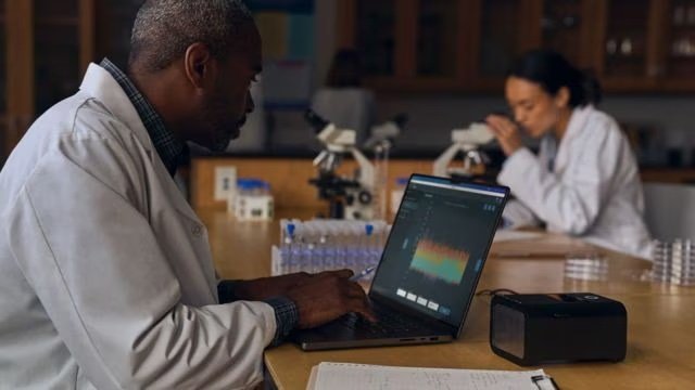 Two scientists in lab coats work at a table with a laptop and microscopes, surrounded by lab equipment and glassware.