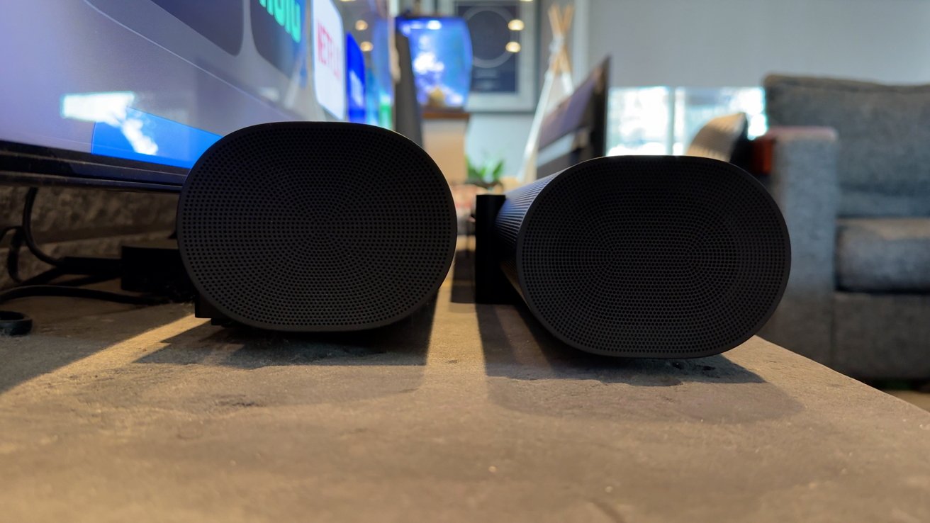 Two black cylindrical speakers side by side on a table in a living room, with a television and sofa in the background.