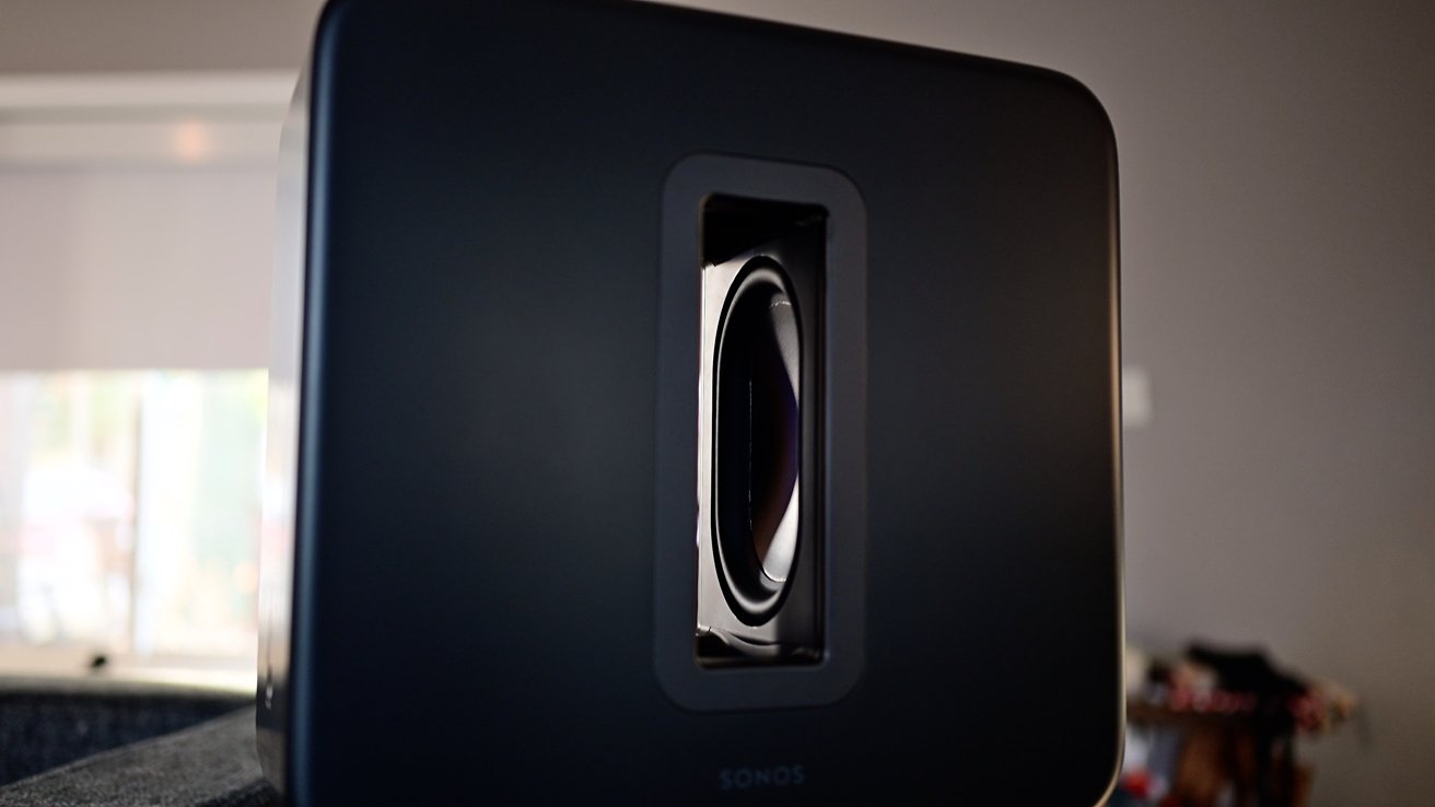 Close-up of a black rectangular speaker with a visible inner woofer, positioned inside a modern room setting.