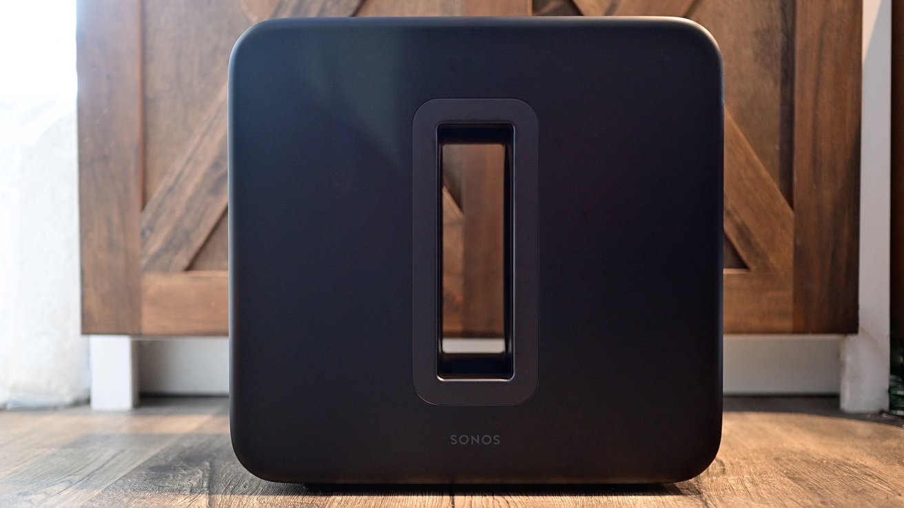 A black Sonos subwoofer with a rectangular opening in the center, placed on a wooden floor in front of a wooden cabinet.