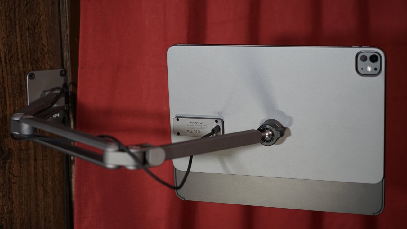 Tablet on a wall-mounted, adjustable arm with a magnetic charging stand against a red curtain background.