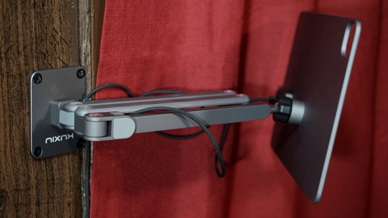 A metallic adjustable wall mount holding a sleek tablet, with a visible cable, against a wooden surface and red curtain backdrop.