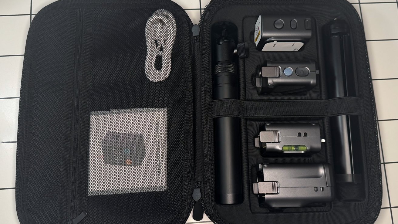 A black protective case holds an organized set of black electronic devices, a white cable, and a small mesh pouch with a manual.