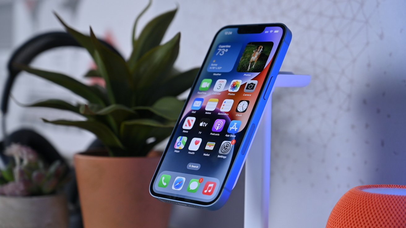 Smartphone on a stand displaying apps with a plant and an orange speaker in the background.