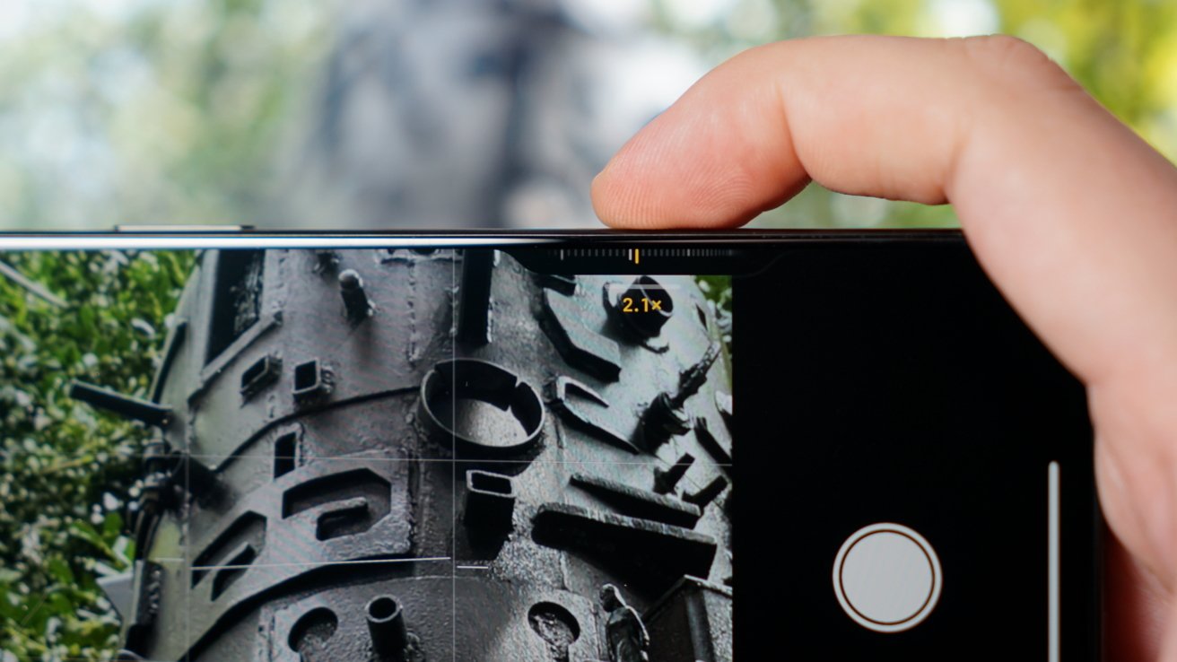 A hand holds a smartphone, capturing a close-up view of a textured, metal sculpture outdoors with greenery in the background.