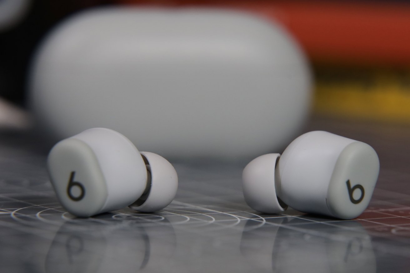 Two white wireless earbuds with a black lowercase 'b' logo, placed on a surface with circular patterns, blurred background featuring a larger white object.