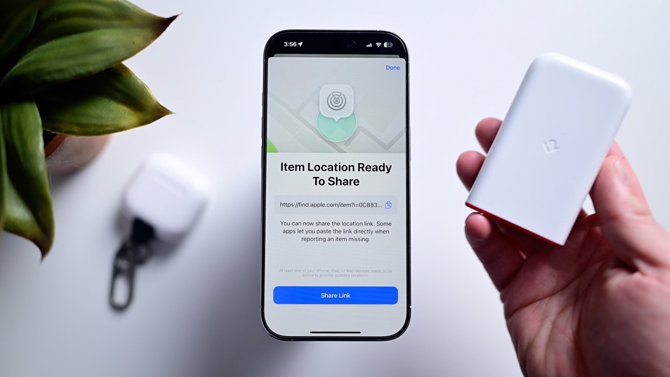 Smartphone displaying item location sharing screen, hand holding a small white device, blurred white AirPods case, and a green potted plant on a white surface.