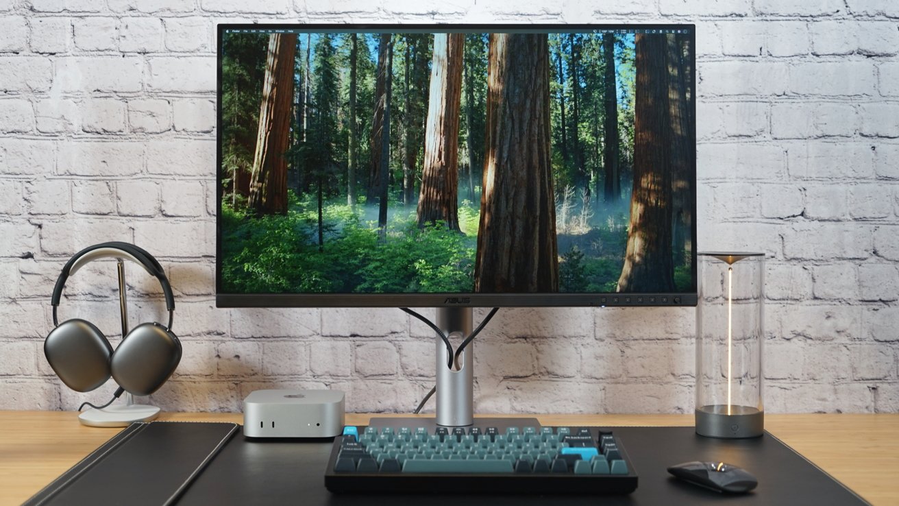 Desktop setup with a monitor displaying a forest scene, headphones on a stand, a mini computer, keyboard, mouse, and hourglass against a brick wall background.