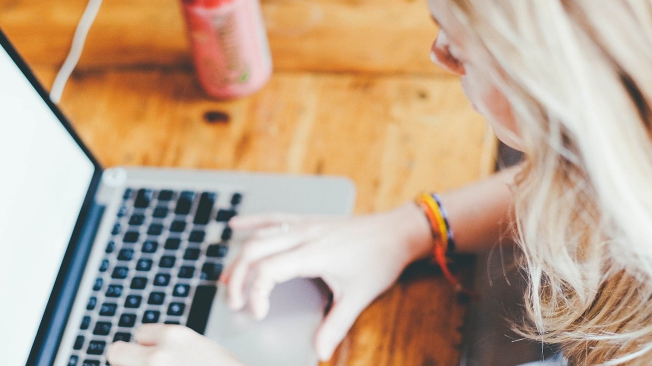 Person using a laptop at a wooden table with a pink drink nearby, hair partially visible.