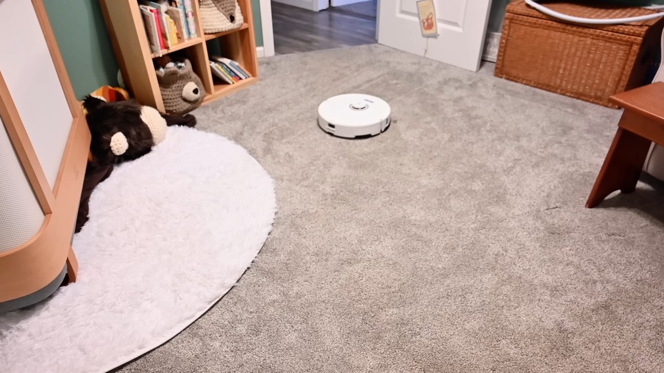 Cozy room with gray carpet, circular white rug, stuffed toys, books on shelves, and a robotic vacuum cleaner on the floor.