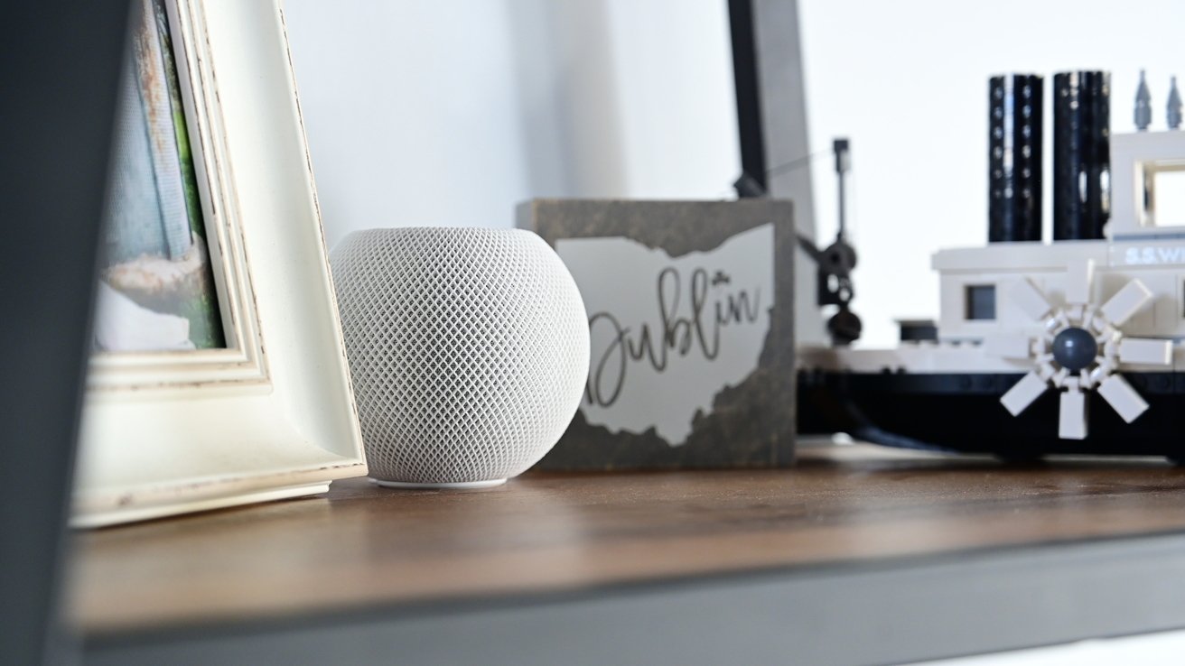 White smart speaker on a wooden shelf next to a framed photo, a Dublin sign, and a model ship.